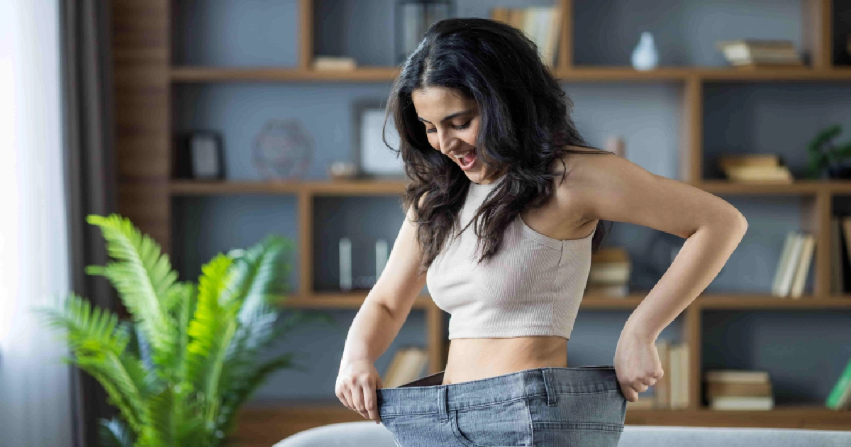 A joyful young woman shows off her oversized jeans, celebrating weight loss with GLP-1 Weight Loss in Chesterfield, MO, with a wide smile in a cozy living room